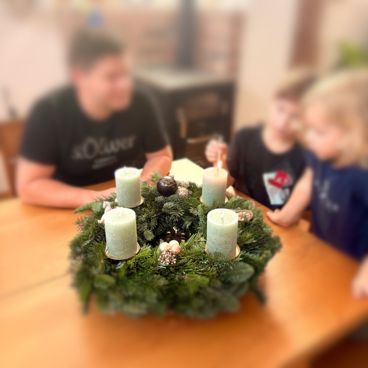 Eine Familie sitzt an einem Tisch mit einem Kranz aus grünen Zweigen und vier brennenden Kerzen. Der Vater trägt ein schwarzes T-Shirt, während die Kinder schwarze und blaue T-Shirts tragen.