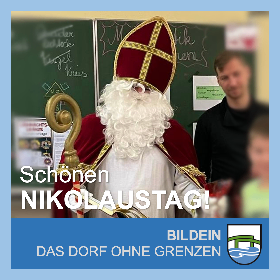 Ein Mann als St. Nikolaus verkleidet steht in einem Klassenzimmer mit einem goldenen Stab und einem Papier in der Hand. Ein Mann und ein Kind sind im Hintergrund.