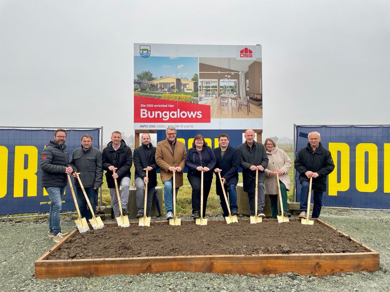 Gruppe von Menschen an einer Baustelle mit Schaufeln. Ein Schild steht 'Bungalows'.