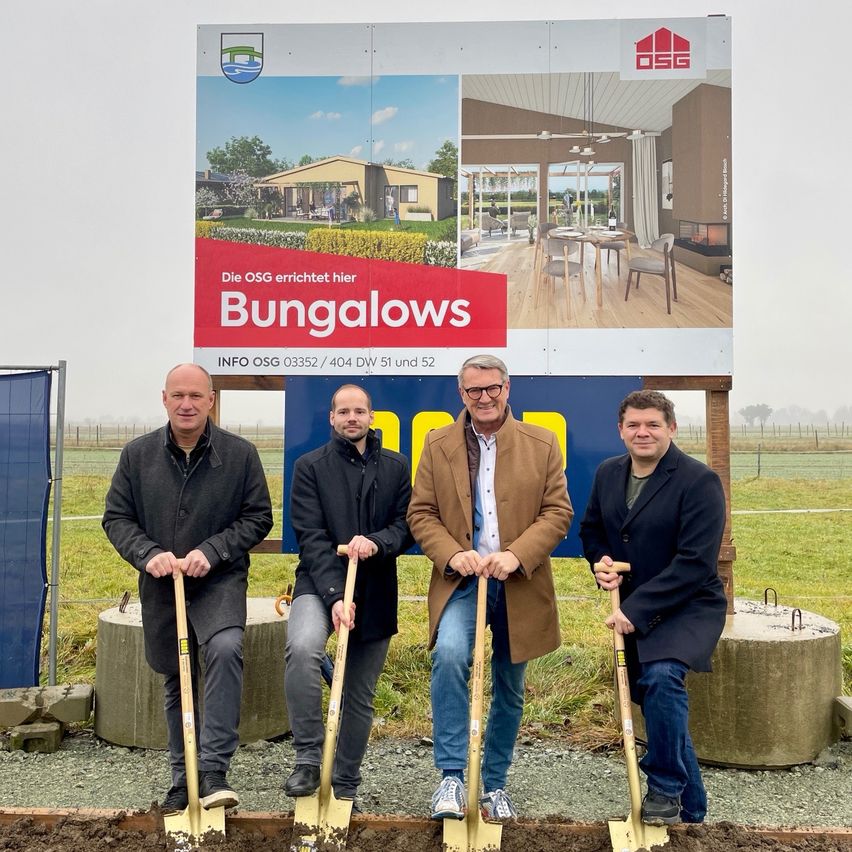 Vier Männer halten Schaufeln vor einem Werbeschild für Bungalows. Hinter ihnen ist ein Grasfeld.