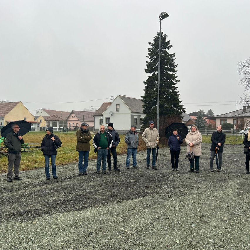 Eine Gruppe von Menschen steht auf einer Kiesstraße und hält Regenschirme. Sie tragen Winterkleidung und scheinen auf etwas zu warten. Hinter ihnen befinden sich Häuser und eine hohe Tanne.