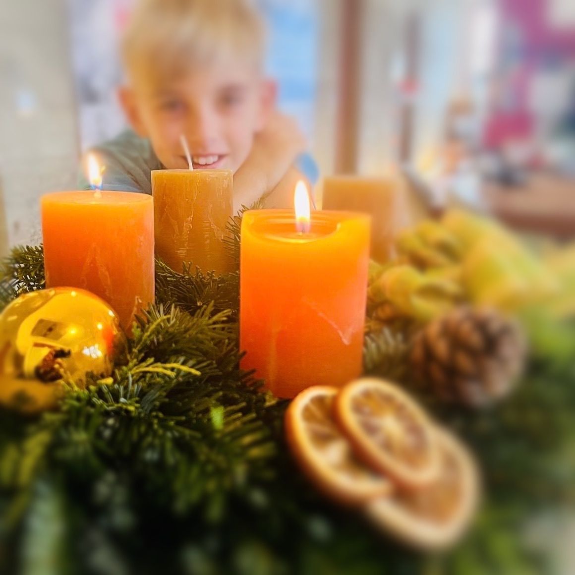 Ein Junge sitzt lächelnd neben Kerzen, Tannenzapfen und Orangen auf einem Tisch.