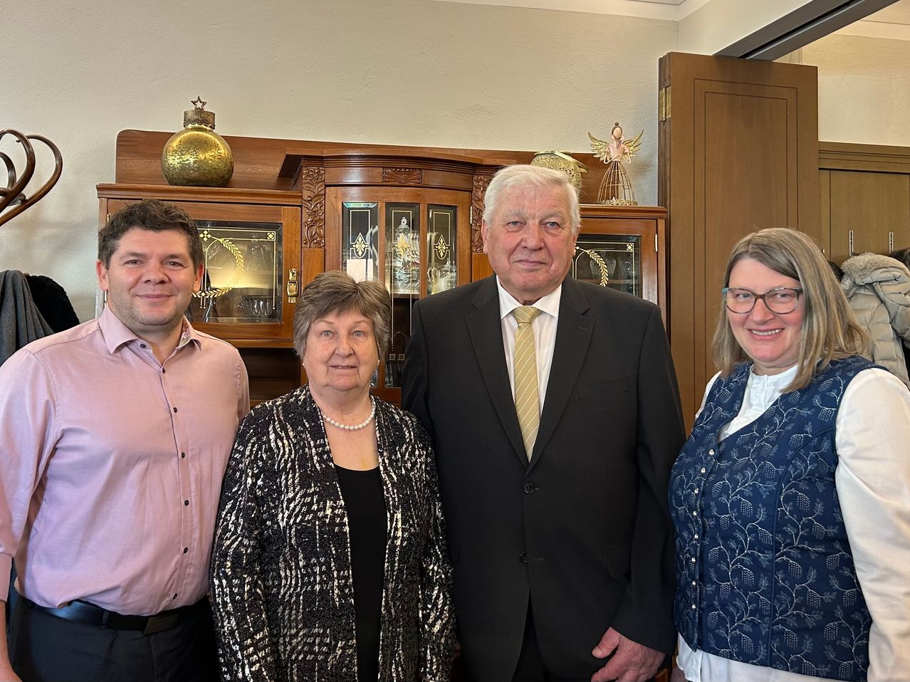 Vier Menschen, zwei Männer und zwei Frauen, stehen eng beieinander vor einem hölzernen Schrank mit Glastüren und lächeln in die Kamera.