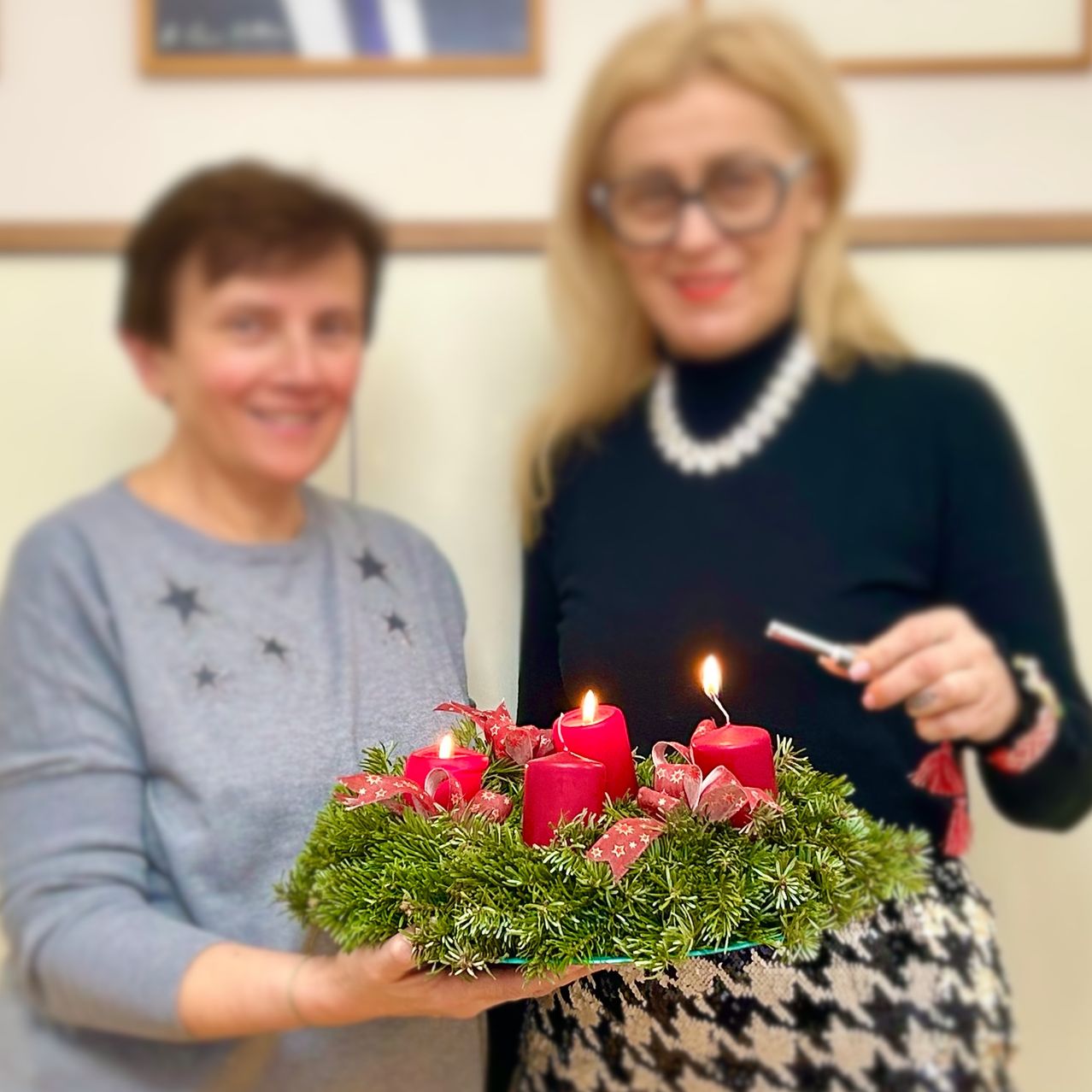 Zwei Frauen lächeln und halten eine Girlande mit vier roten Kerzen und roten Bändern. Eine Frau hält ein Feuerzeug.