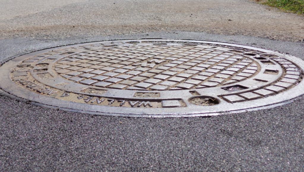 Ein kreisförmiger Metall-Mannlochdeckel mit Kreuzmuster auf der Oberfläche. Er hat eine silberne Farbe und ist auf der Straße platziert.
