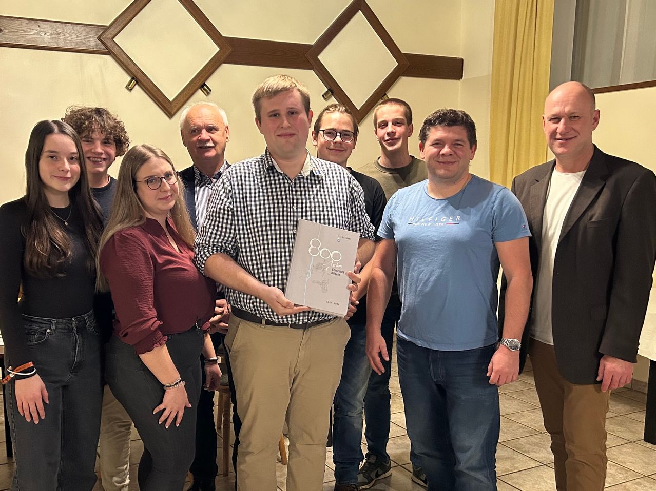 Eine Gruppe von Menschen posiert für ein Foto mit einem Mann, der ein Buch hält. Hinter ihm stehen zwei Männer mit Brille und eine Frau mit Armband. Rechts ist ein Mann, der eine Uhr trägt. Im Hintergrund ist eine Wand mit einem Kreuzdesign.