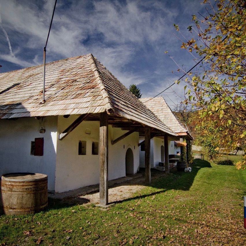 Ein altes rustikales Haus mit einem Ziegeldach und Holzsäulen steht in einem Rasen, mit einem Fass in der Nähe.