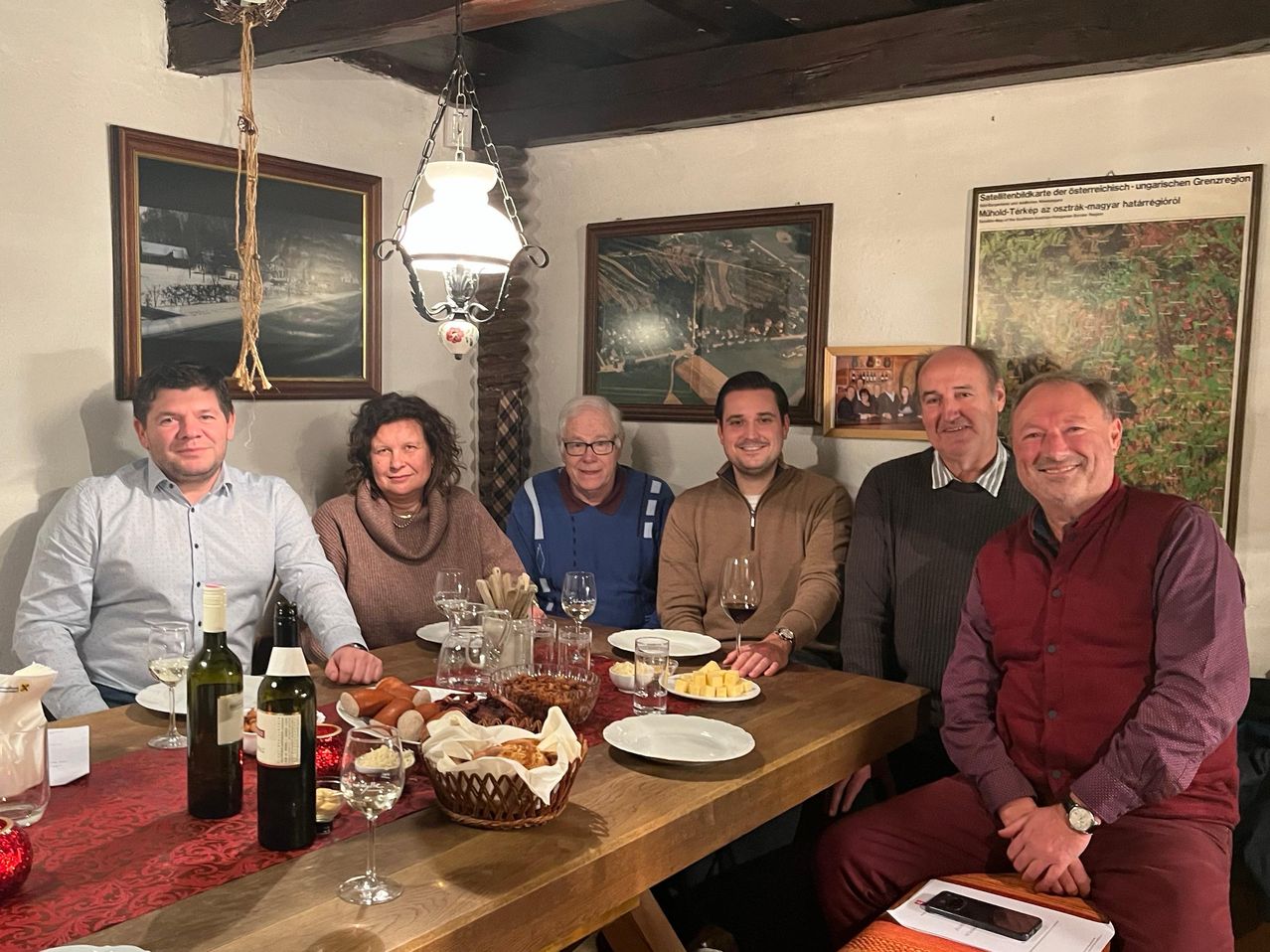 Eine Gruppe von sechs Personen sitzt um einen Esstisch herum mit verschiedenen Speisen, Weingläsern und Flaschen. An der Wand hinter ihnen hängen Gemälde.