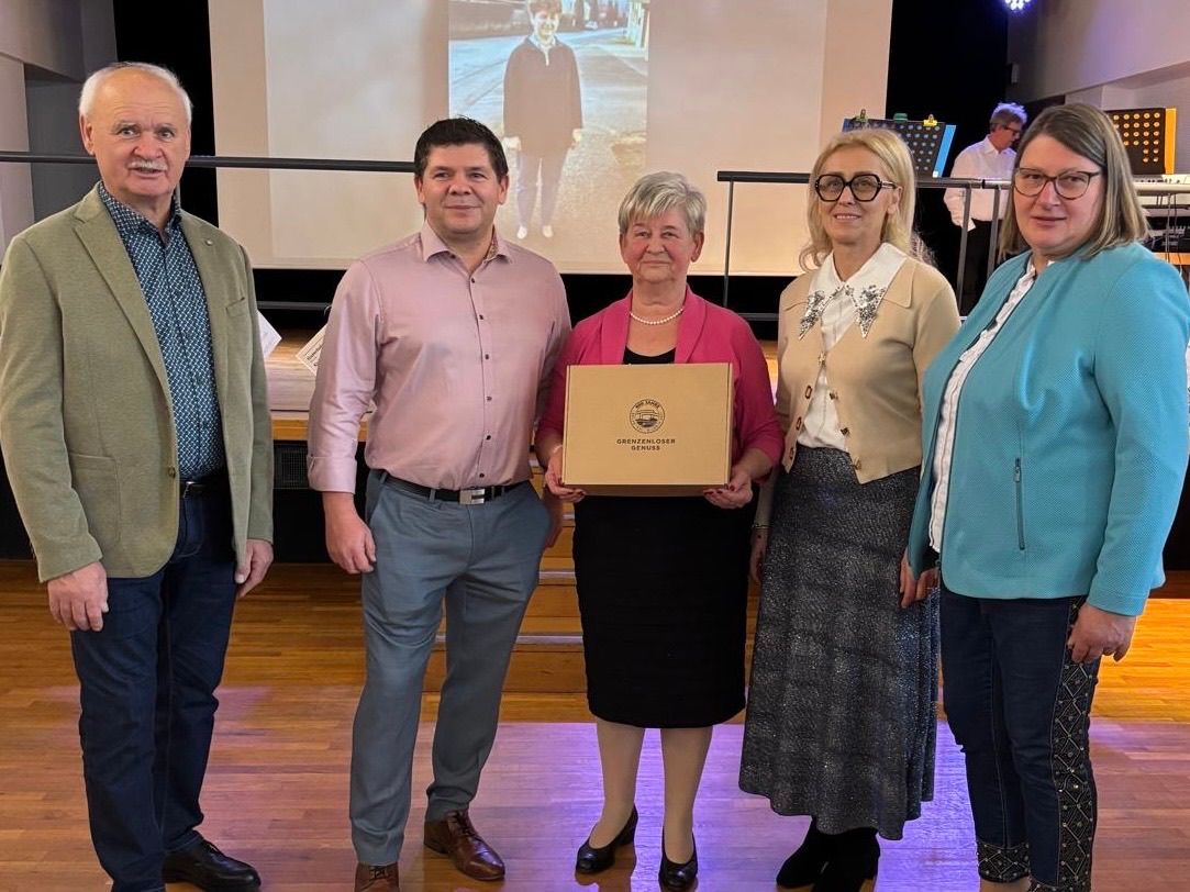 Fünf Personen stehen auf einer Bühne, eine Frau hält ein Zertifikat und lächelt. Dahinter zeigt eine Leinwand ein Bild einer Person, die geht.