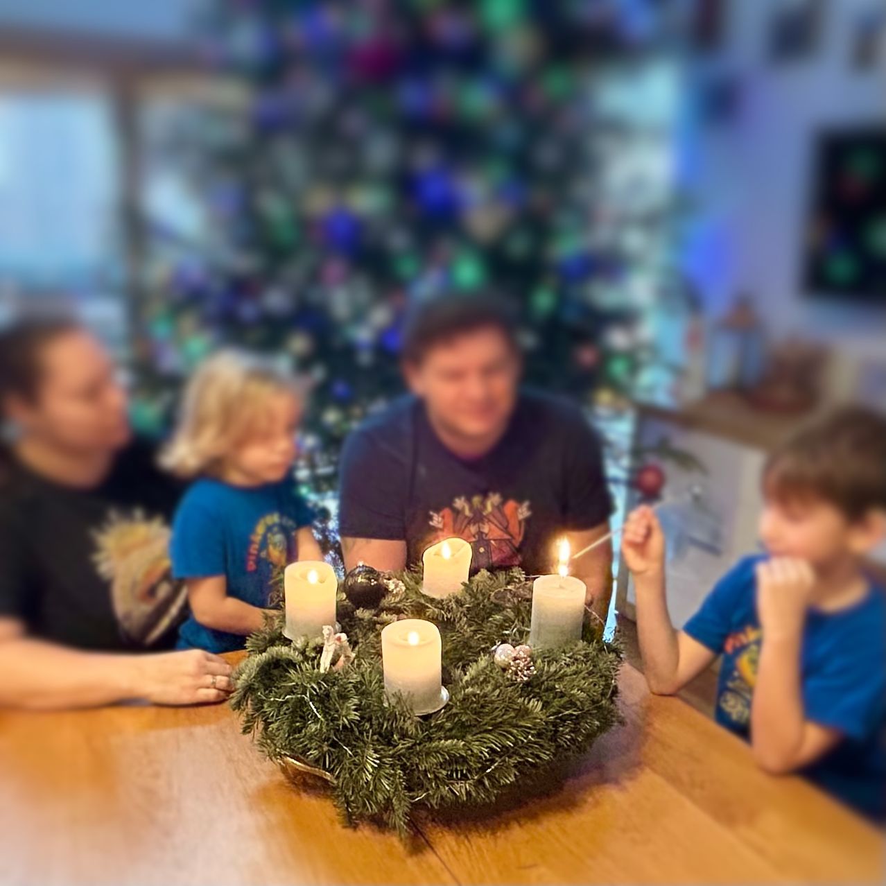 Eine Familie versammelt sich um einen Tisch mit einem Kranz aus brennenden Kerzen und lächelt vor einem geschmückten Weihnachtsbaum.