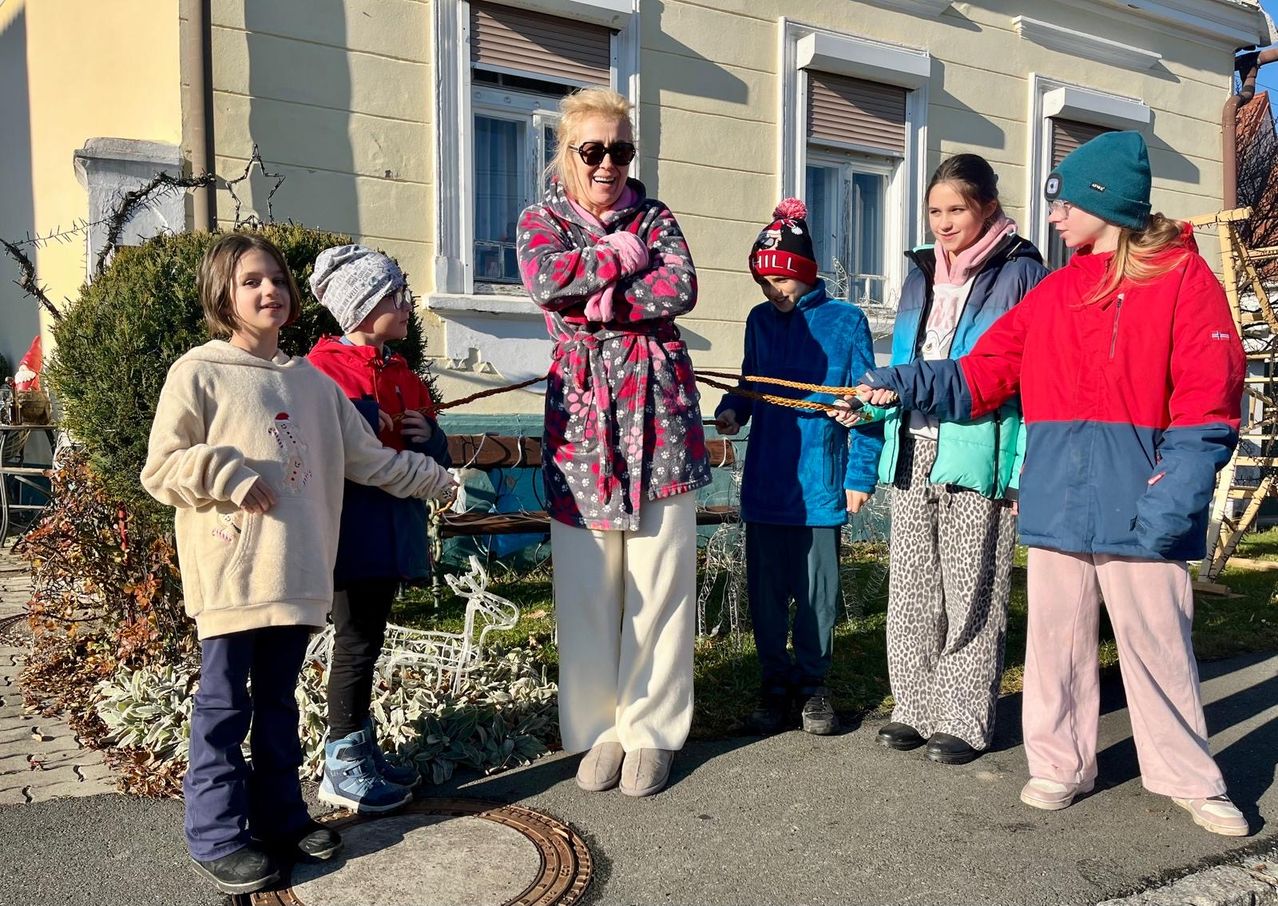 Eine Frau und sechs Kinder stehen vor einem Gebäude. Die Frau lächelt und hält zwei Seile. Die Kinder stehen um sie herum, lächeln und halten Seile.
