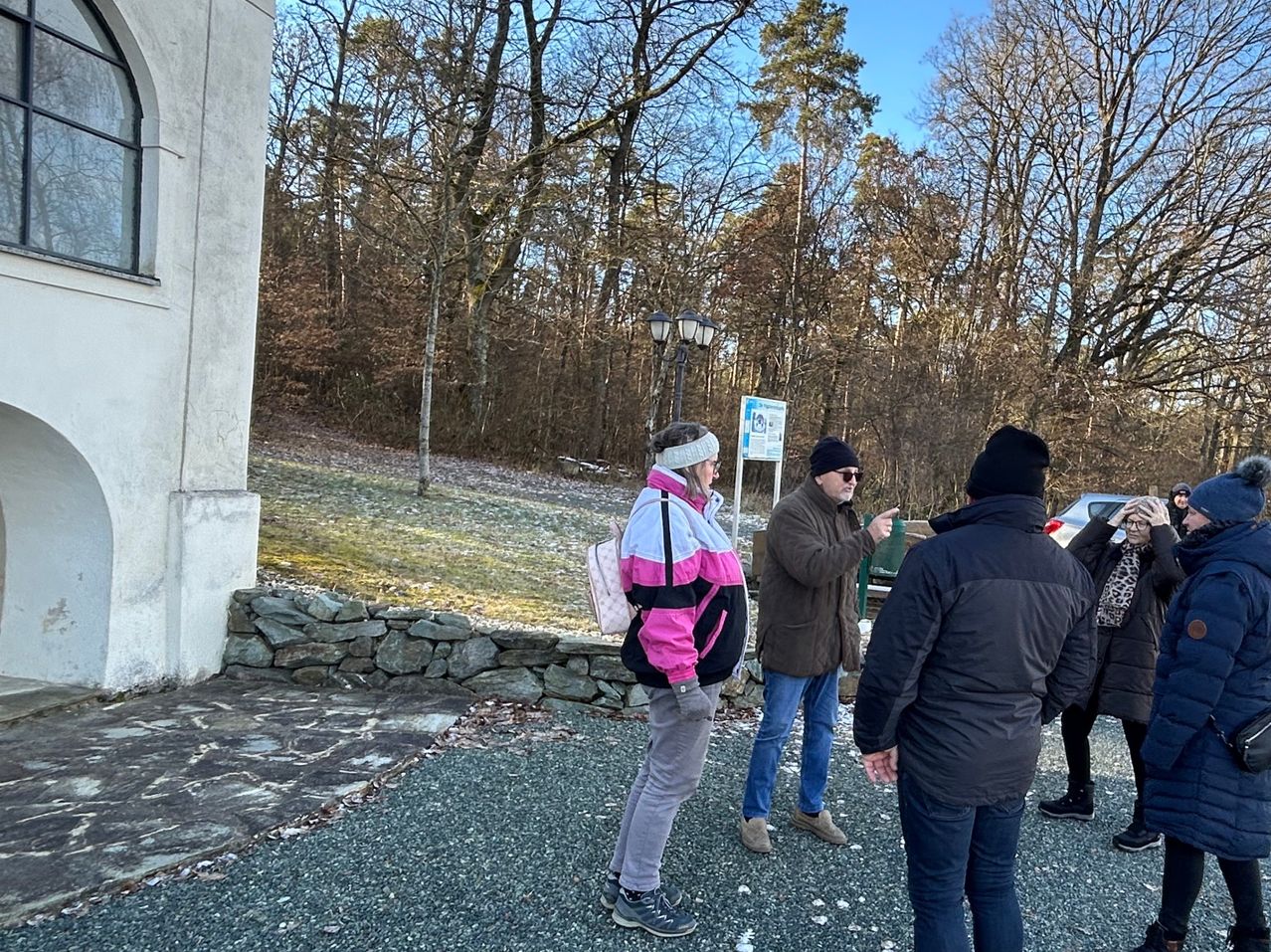 Drei Personen stehen in einem Außenbereich und besprechen etwas. Eine Person links trägt eine pinke Jacke. Ein anderer Mann in einer braunen Jacke zeigt auf etwas und spricht. Der Himmel ist klar.