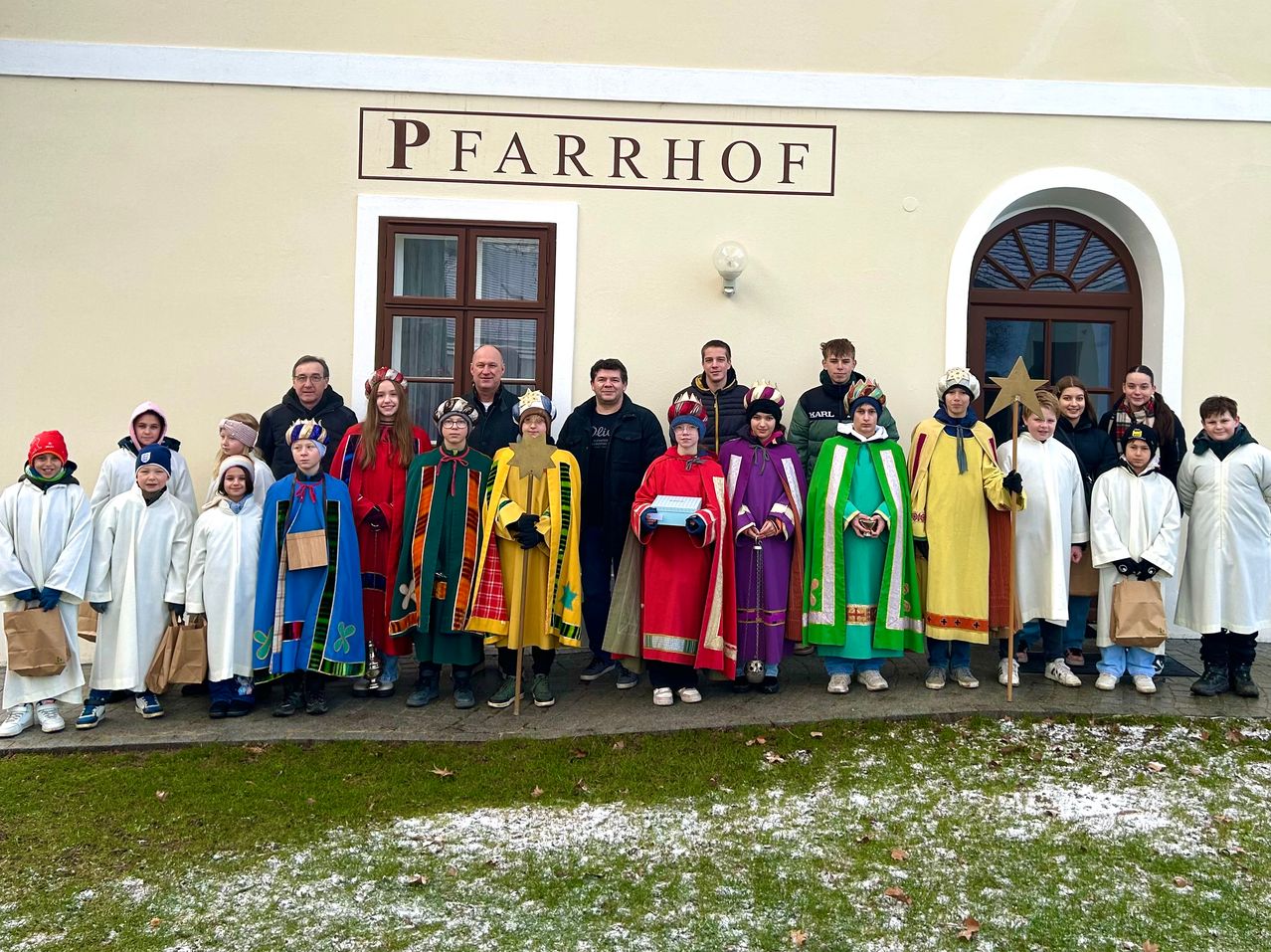 Eine Gruppe von Menschen in bunten Kostümen posiert für ein Foto vor dem Pfarrhof-Gebäude. Einige tragen Hüte, während andere einen Stern halten.