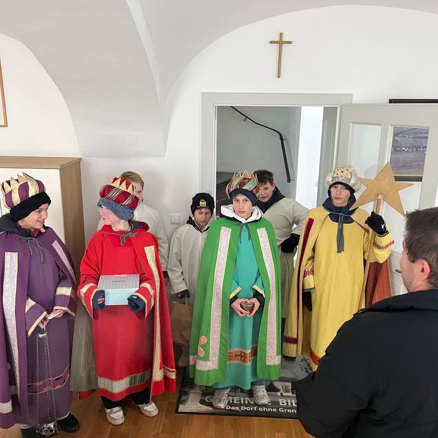 Eine Gruppe von Menschen in mittelalterlichen Kostümen steht in einem Raum mit gewölbten Decken. Sie tragen Kronen und halten verschiedene Gegenstände, darunter einen Stern und eine Schachtel. Ein Mann macht ein Foto von ihnen.