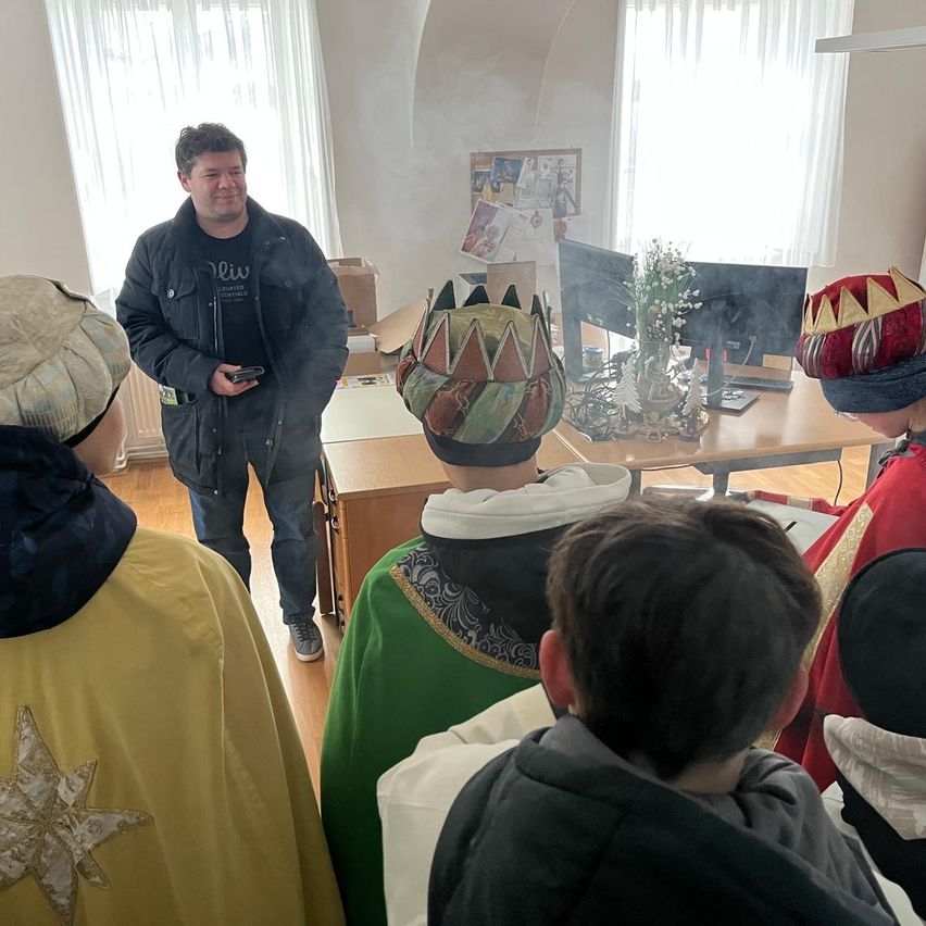 Ein Mann steht vor einem Schreibtisch mit Monitoren und spricht zu einer Gruppe von Kindern in Kostümen. Der Raum hat weiße Wände, Holzböden und ein Fenster mit Jalousien.
