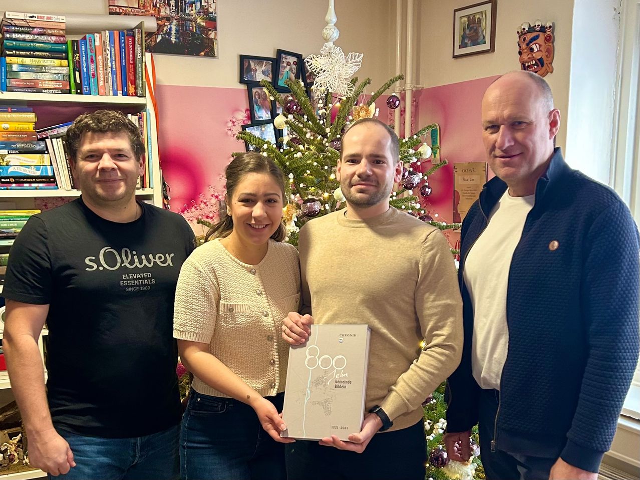 Vier Personen stehen zusammen vor einem geschmückten Weihnachtsbaum. Ein Mann rechts hält ein Buch, während eine Frau in der Mitte in die Kamera lächelt. Ein Mann links trägt ein schwarzes Shirt.