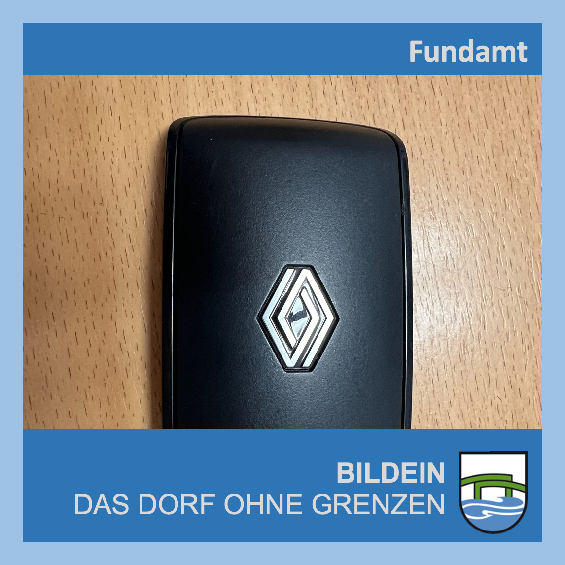 Ein schwarzes rechteckiges Objekt mit einem rautenförmigen Renault-Logo liegt auf einer Holzoberfläche. Das Bild enthält Textüberlagerungen 'Fundamt' und 'Bildein das Dorf ohne Grenzen.' Ein Emblem mit einem welligen Wasserdesign ist ebenfalls vorhanden.