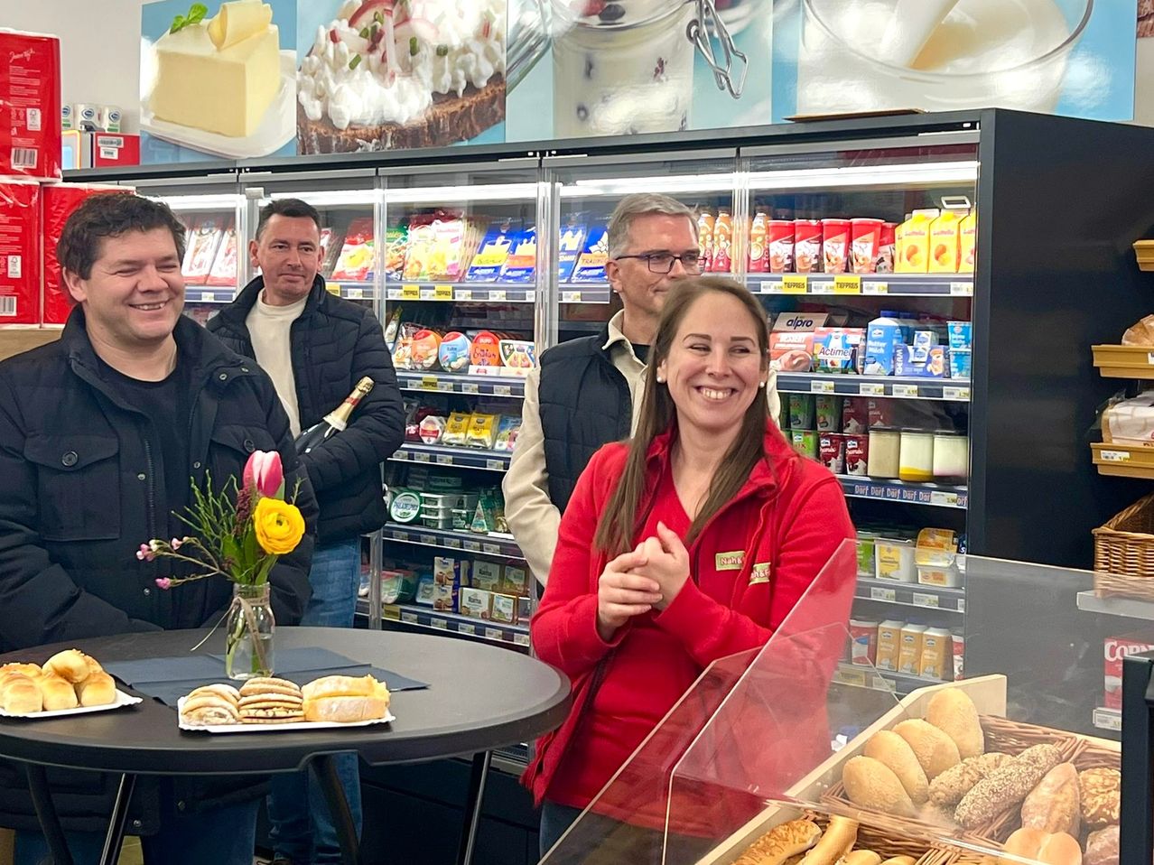 Vier Menschen stehen lächelnd in einem Lebensmittelgeschäft. Eine Frau in einer roten Jacke steht neben einem Tisch mit einem Blumenstrauß und Brot. Dahinter befindet sich eine Vitrine mit verschiedenen Lebensmitteln.