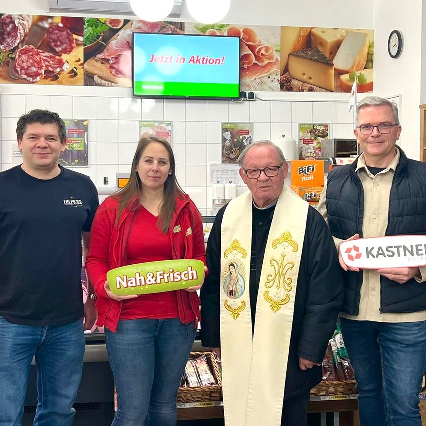 Vier Personen posieren für ein Foto in einem Geschäft. Eine Frau hält ein Schild mit der Aufschrift 'Nah&Frisch'. Ein Mann in einem weißen und goldenen Gewand steht zwischen zwei Männern in Jacken. Hinter ihnen zeigt ein Monitor 'Jetzt in Aktion!' mit verschiedenen Lebensmitteln im Hintergrund.