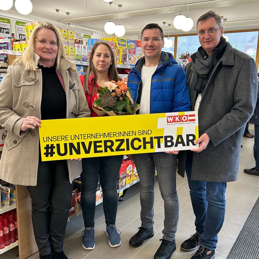 Vier Personen in Winterkleidung stehen in einem Geschäft und halten ein gelbes Schild. Das Schild trägt die Aufschrift 'Unsere UnternehmerInnen sind #unverzichtbar' und wird von den beiden Männern gehalten. Hinter ihnen sind Regale mit Produkten zu sehen.