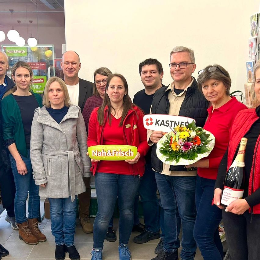 Eine Gruppe von Menschen steht in einem Raum zusammen. Eine Frau in Rot hält ein Schild mit der Aufschrift Nah&Frisch. Eine andere Frau hält einen Blumenstrauß. Sie alle lächeln und scheinen für ein Foto zu posieren.