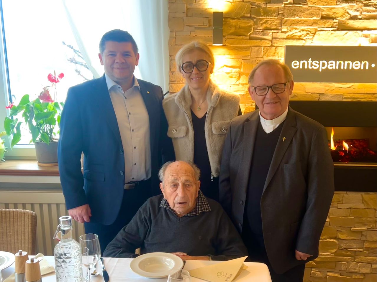 Vier Menschen stehen und sitzen zusammen in einem Raum. Ein älterer Mann sitzt an einem Tisch, während ein Mann und eine Frau hinter ihm stehen. Ein weiterer Mann steht rechts. Ein Schild mit der Aufschrift 'entspa' befindet sich an der Wand.