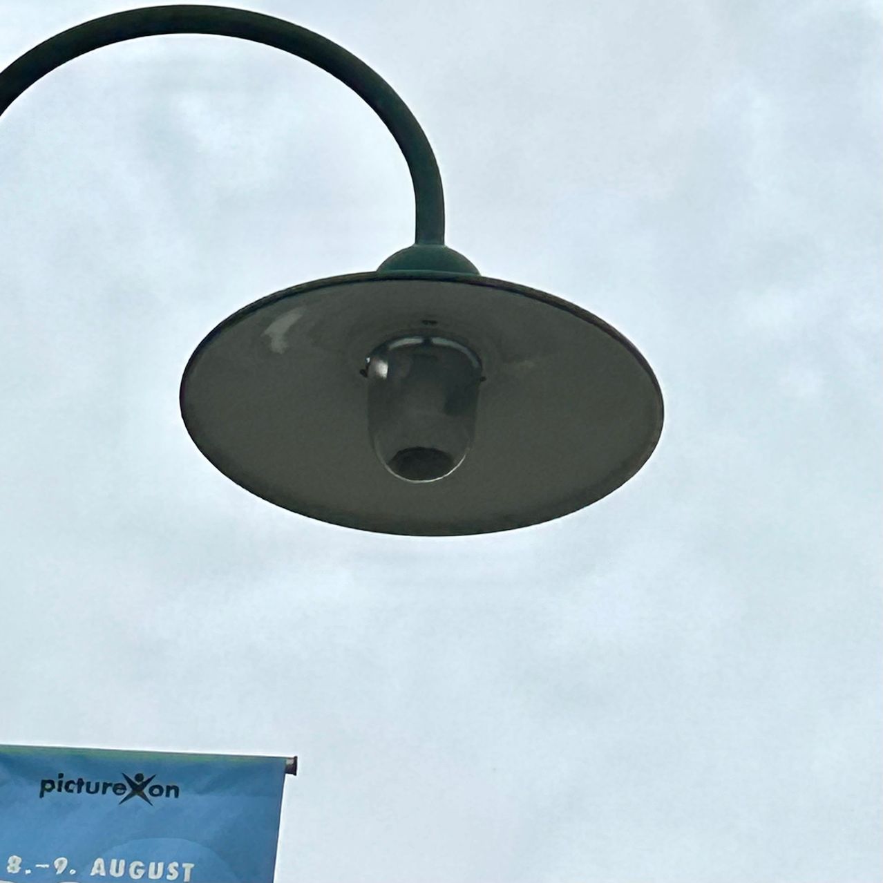 Eine Straßenlampe mit einem gebogenen Arm und einer kreisförmigen Leuchte hängt unter einem bewölkten Himmel. Darunter steht ein blauer Banner mit PictureXon und 8.-9. August.
