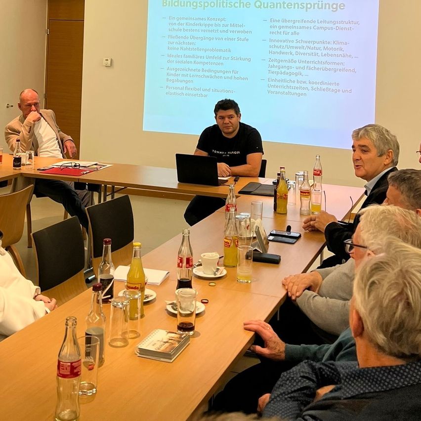 Eine Gruppe von Personen sitzt in einem Besprechungsraum um einen Tisch herum und führt eine Diskussion. Ein Laptop und eine Projektionsleinwand mit dem Titel 'Bildungspolitische Quantensprünge' sind sichtbar. Flaschen und andere Gegenstände befinden sich auf dem Tisch.