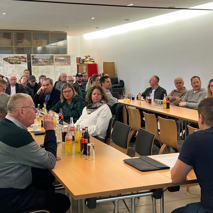 Eine Gruppe von Menschen sitzt in einem Konferenzraum an Tischen, mit Getränken und Snacks auf den Tischen. Einige Personen führen Gespräche, während andere auf ihren Laptops zu arbeiten scheinen. Der Raum ist gut beleuchtet mit Deckenlampen.