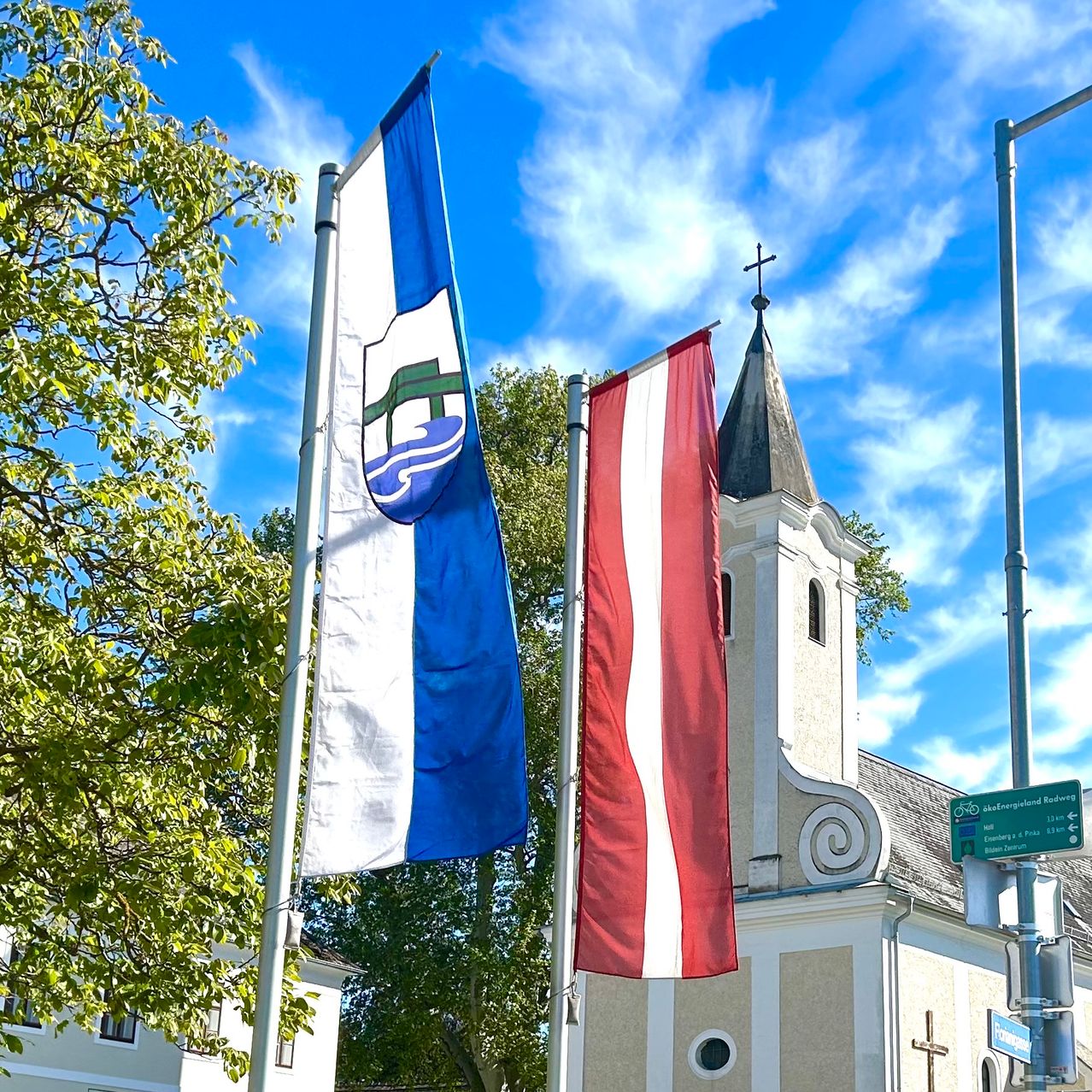 A church with a steeple and a cross is in front of a blue and white flag. Trees and a green signboard are nearby.