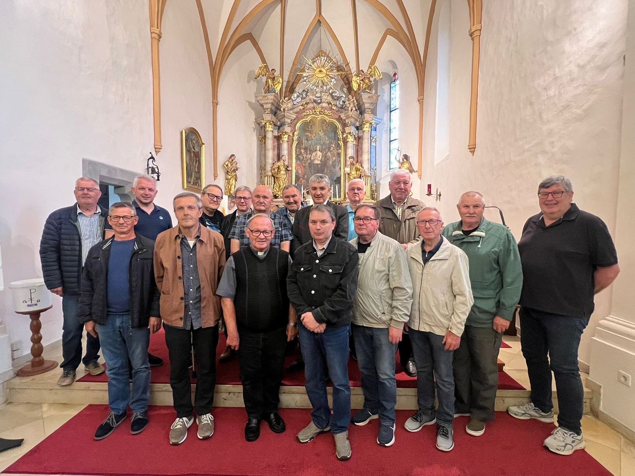 Eine Gruppe von Männern, einige mit Brille, stehen in einer Kirche mit goldenem Altar und kunstvollen Gemälden. Sie posieren für ein Foto auf einem roten Teppich.