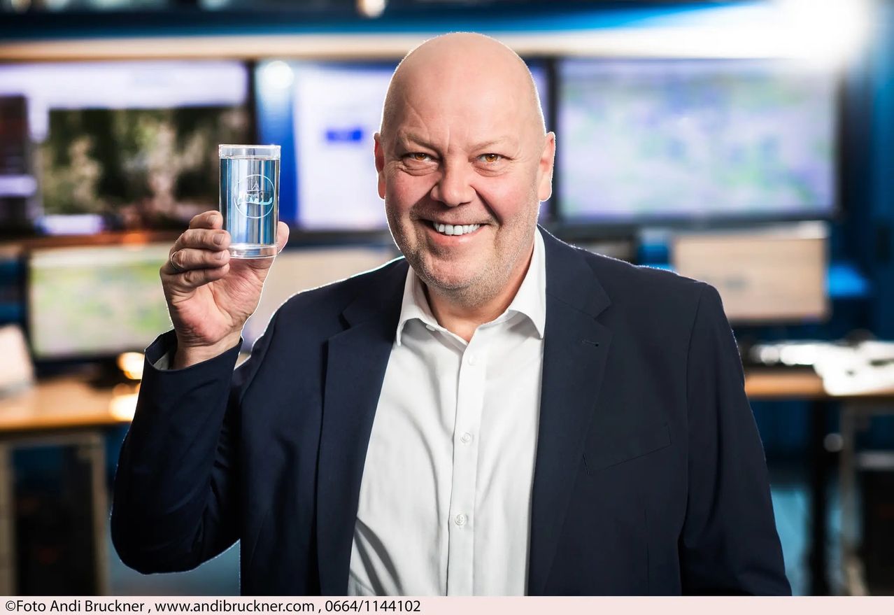 Ein Mann in einem Anzug hält ein Glas Wasser und lächelt in die Kamera, in einem Kontrollraum mit Monitoren hinter ihm.