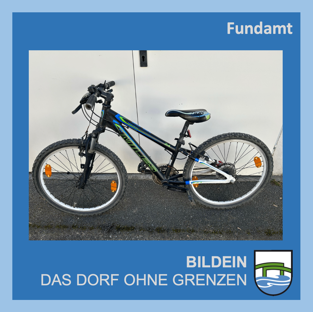 Ein schwarz-blaues Fahrrad mit orangefarbenen Reflektoren steht vor einer weißen Wand. Darunter ist der Text 'Das Dorf ohne Grenzen' zu sehen, zusammen mit einem Logo.