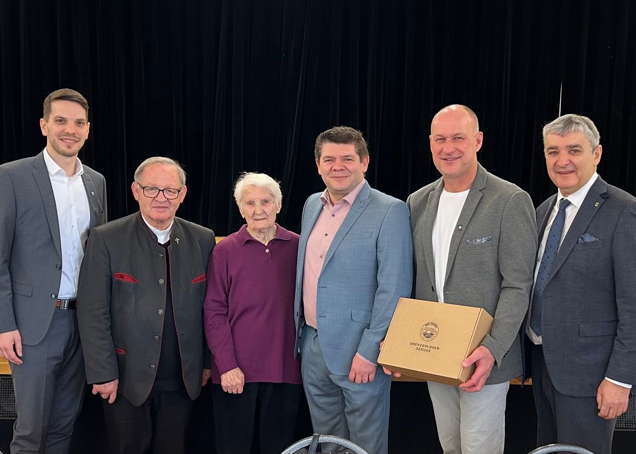 Fünf Personen stehen zusammen in einer formellen Umgebung und lächeln. Eine Frau in einem lila Pullover steht zwischen zwei Männern, einer hält eine Schachtel mit der Aufschrift Grenzloser Genuss. Hinter ihnen ist ein schwarzer Vorhang.