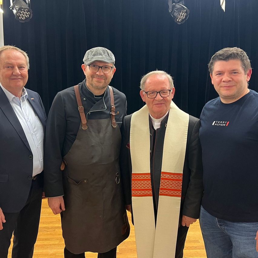 Vier Männer stehen auf einem Holzboden und lächeln für ein Foto. Sie tragen formelle Kleidung und einer der Männer trägt eine Schürze. Hinter ihnen befindet sich ein schwarzer Vorhang mit Scheinwerfern.