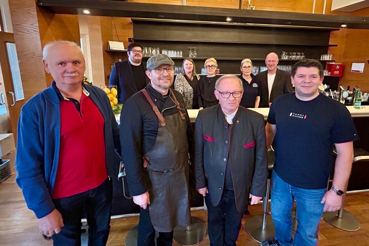 Eine Gruppe von Menschen, darunter Köche und Geschäftsleute, steht in einer Küche. Sie lächeln und scheinen für ein Foto zu posieren. Hinter ihnen ist eine Theke zu sehen.