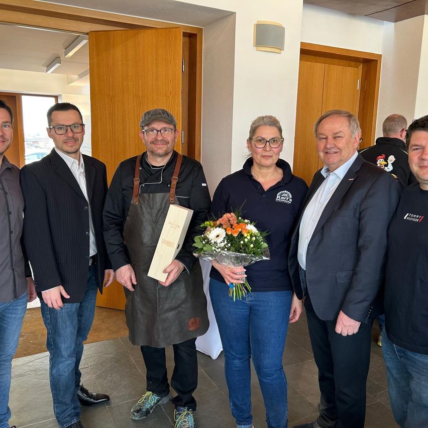 Eine Gruppe von Menschen steht in einem Raum mit Holztüren und einem gefliesten Boden. Die Frau hält einen Blumenstrauß. Der Mann neben ihr trägt eine Lederschürze und hält eine Holzkiste.