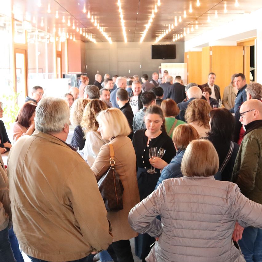 Eine große Gruppe von Menschen ist in einem Raum mit heller Beleuchtung versammelt, einige halten Weingläser. Der Raum hat Glasfenster, Holztüren und einen an der Wand montierten Monitor.