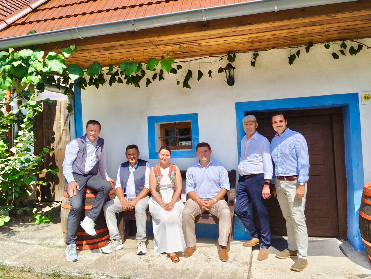 Eine Gruppe von fünf Männern und einer Frau posiert für ein Foto vor einem traditionellen Haus. Die Männer tragen hellblaue Hemden und Hosen, während die Frau ein weißes Kleid trägt. Sie sitzen auf einer Holzbank und stehen auf einem Betonuntergrund.