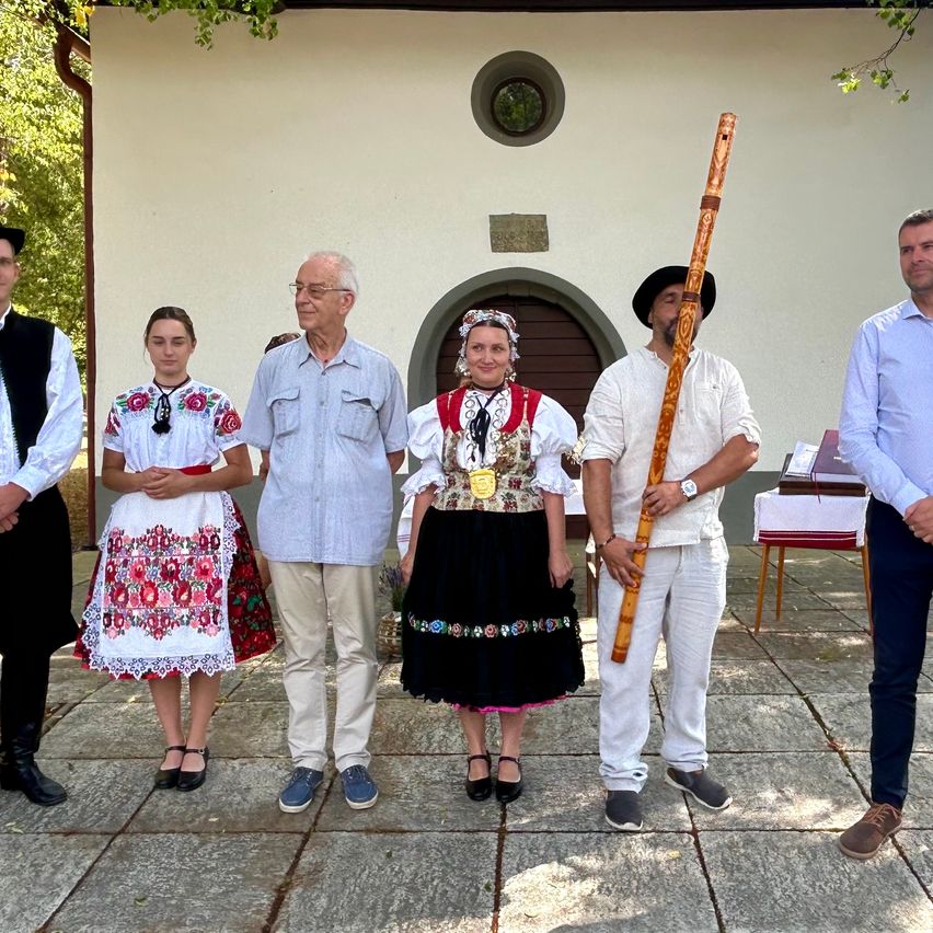 Eine Gruppe von Menschen in traditioneller Kleidung steht vor einem Gebäude. Ein Mann hält eine Holzflöte, und eine Frau in der Mitte trägt eine Medaille. Dahinter ist ein Tisch mit einem Buch zu sehen.