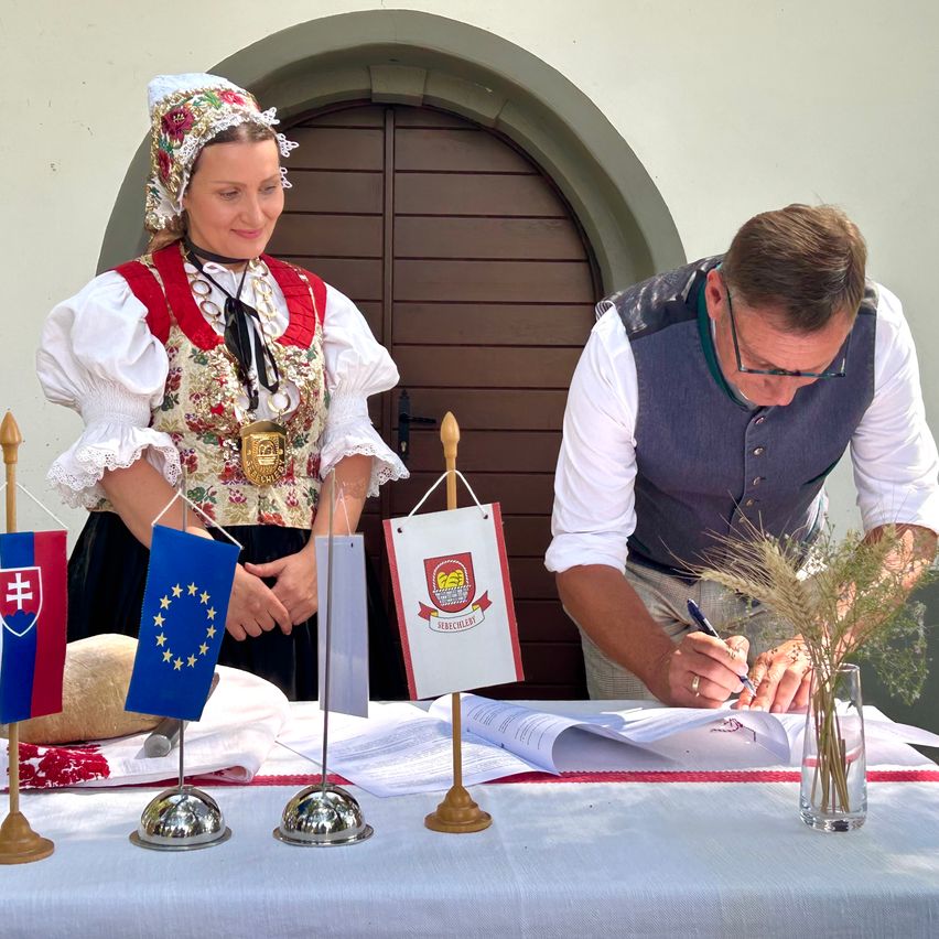 Eine Frau in traditioneller Kleidung steht neben einem Mann in formeller Kleidung, beide unterzeichnen ein Dokument auf einem Tisch, geschmückt mit Flaggen und einem Laib Brot.