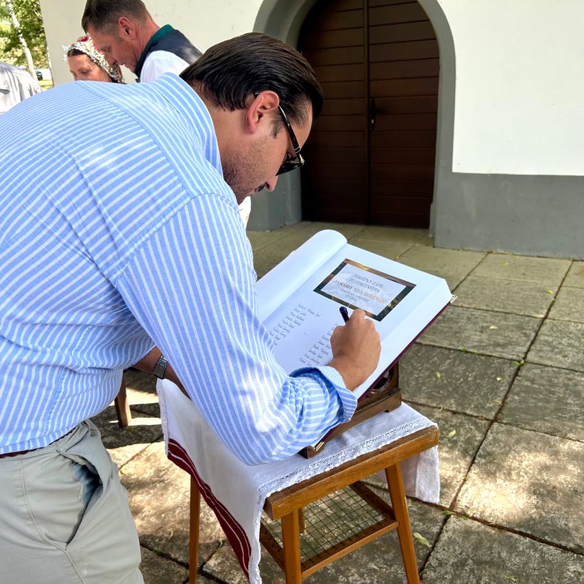 Ein Mann in einem blauen Hemd und mit Brille schreibt in ein Buch auf einem Holzhocker vor einem Gebäude.