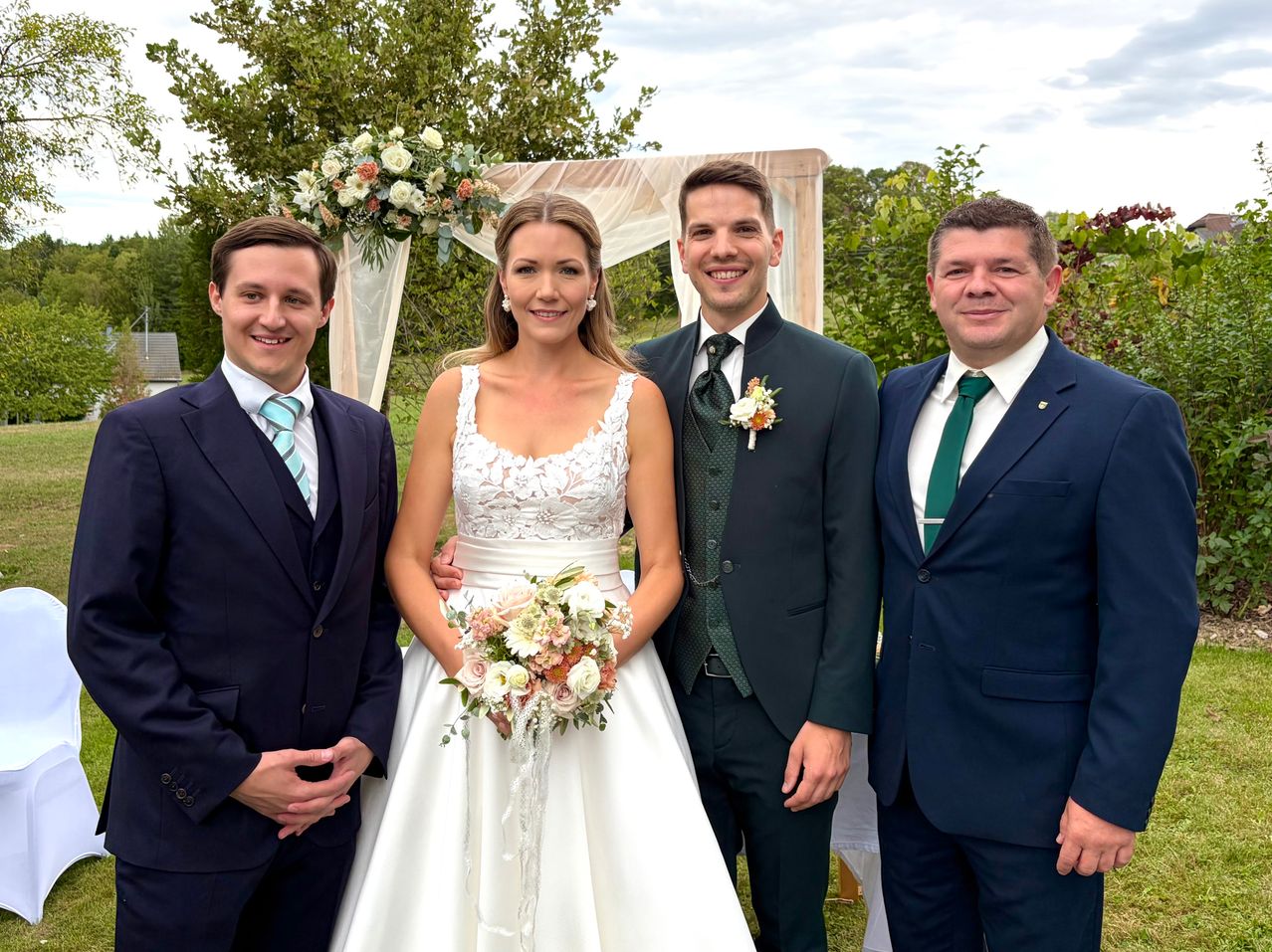 Eine Hochzeitsgesellschaft steht lächelnd zusammen, die Braut in einem weißen Kleid mit einem Blumenstrauß und drei Bräutigams in Anzügen.