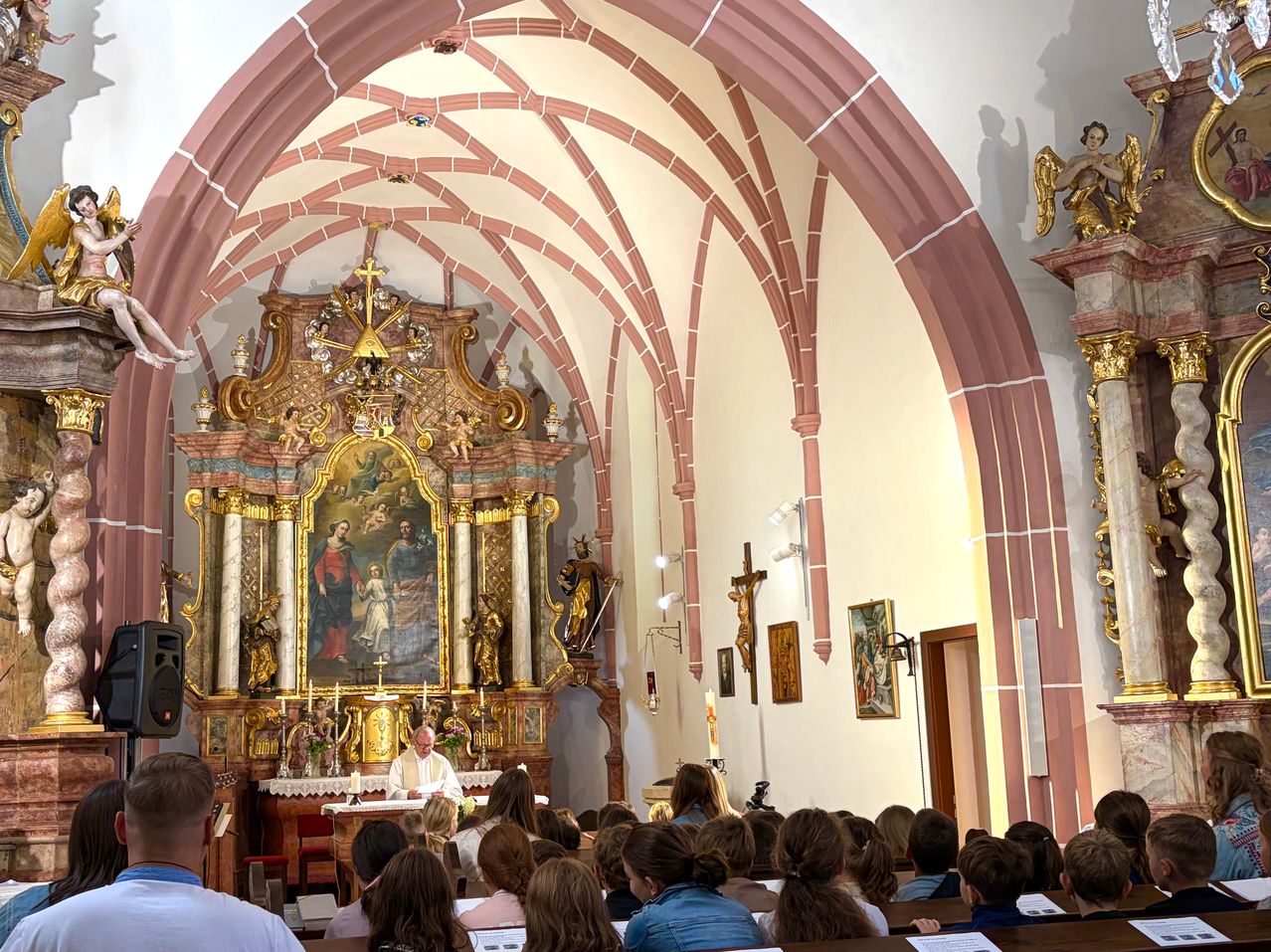 Ein Priester hält eine Predigt in einer Kirche mit einem großen Altarbild hinter ihm. Das Altarbild zeigt eine Darstellung der Jungfrau Maria und Jesu. Die Kirche hat eine hohe Decke mit komplizierten Bögen und ist mit Menschen gefüllt.