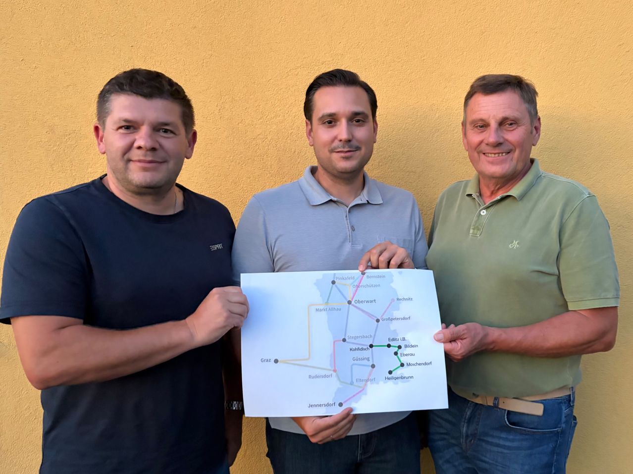 Drei Männer stehen nebeneinander und halten eine Karte mit einer markierten Route in den Händen. Der Mann in der Mitte hält die Karte mit beiden Händen. Der Mann auf der linken Seite trägt ein schwarzes Shirt, und der Mann auf der rechten Seite trägt ein grünes Shirt.
