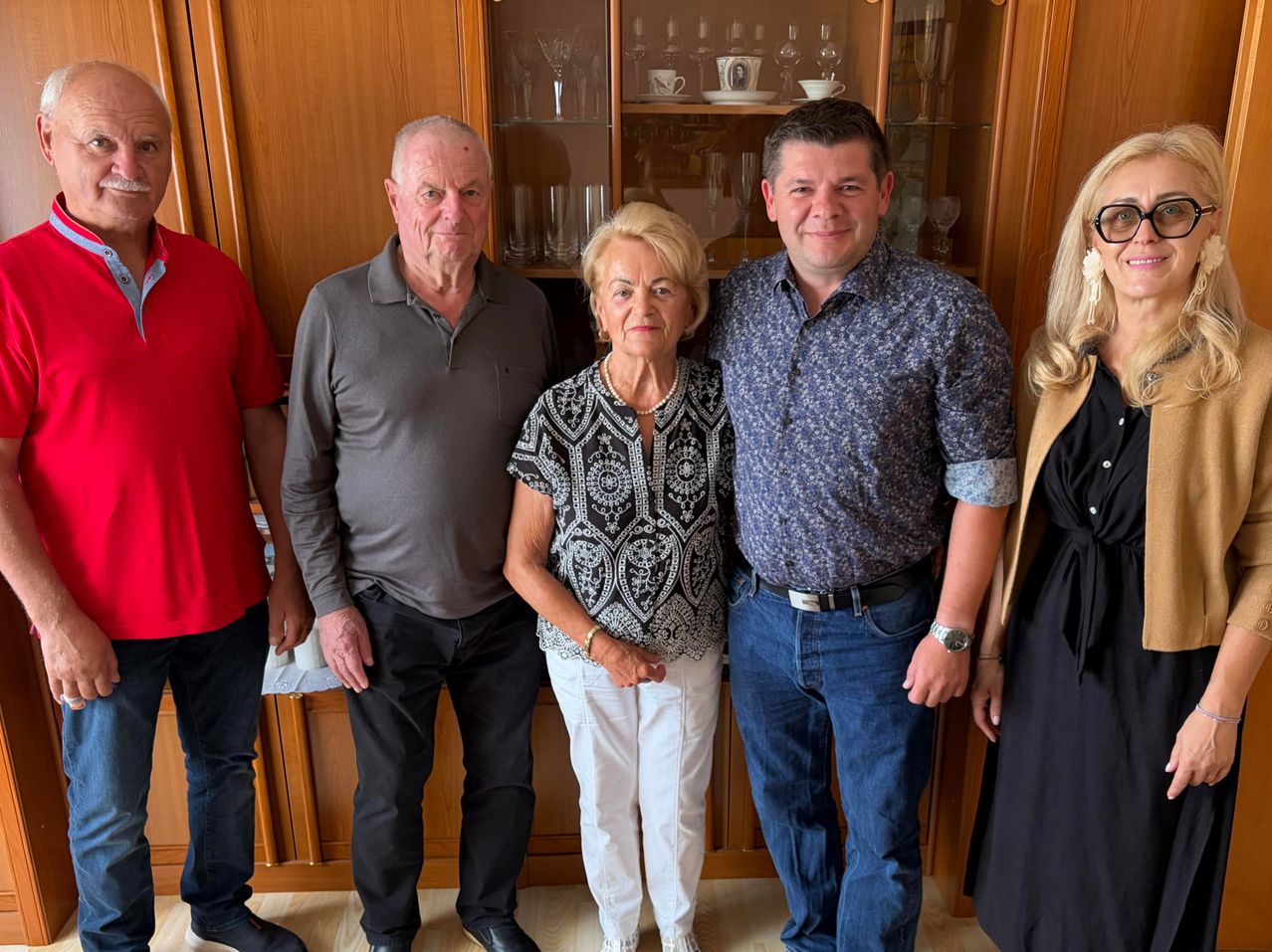 Fünf Menschen stehen vor einem Holzschrank mit Glastüren und lächeln für ein Foto. Der Mann links trägt ein rotes Shirt und der Mann rechts ein schwarzes Kleid. Die Frau in der Mitte trägt eine Halskette und eine Armbanduhr. Der Mann neben ihr trägt eine Uhr.