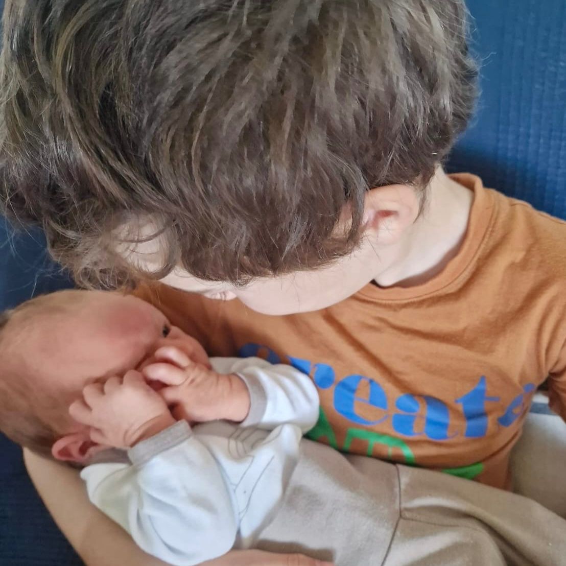 Ein Junge in einem orangefarbenen Shirt hält ein Baby und berührt sanft dessen Gesicht mit seiner Hand. Das Baby schaut ihn an.