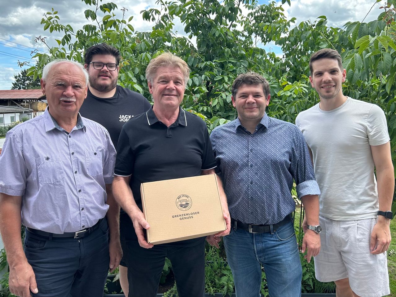 Fünf Männer posieren für ein Foto im Garten, einer hält eine Box. Einer trägt eine Uhr, zwei tragen Gürtel. Hinter ihnen steht ein blattreicher Baum.