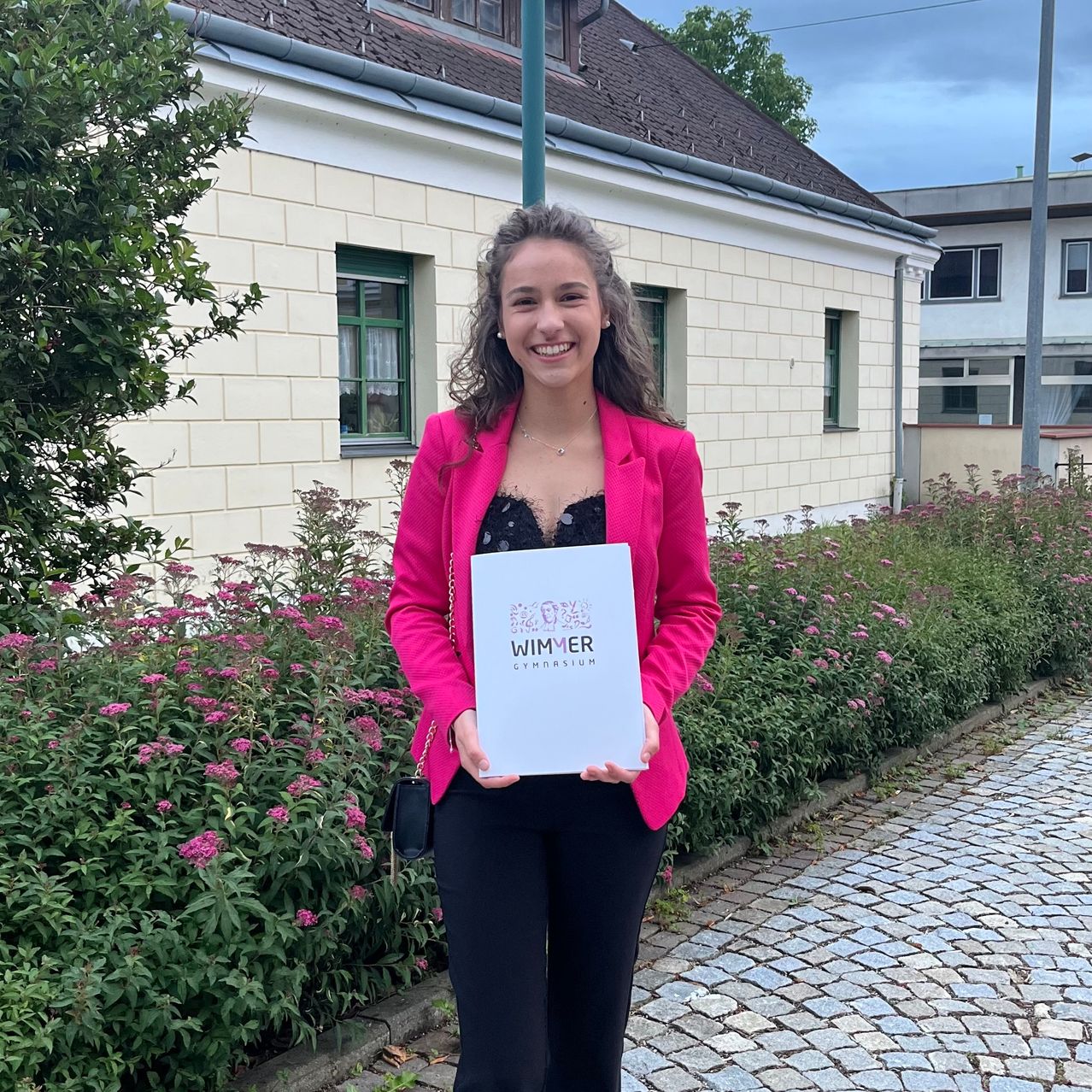 A woman stands outdoors, smiling, holding a white certificate. She wears a pink blazer and black pants. Behind her, a building with green windows and pink flowers.