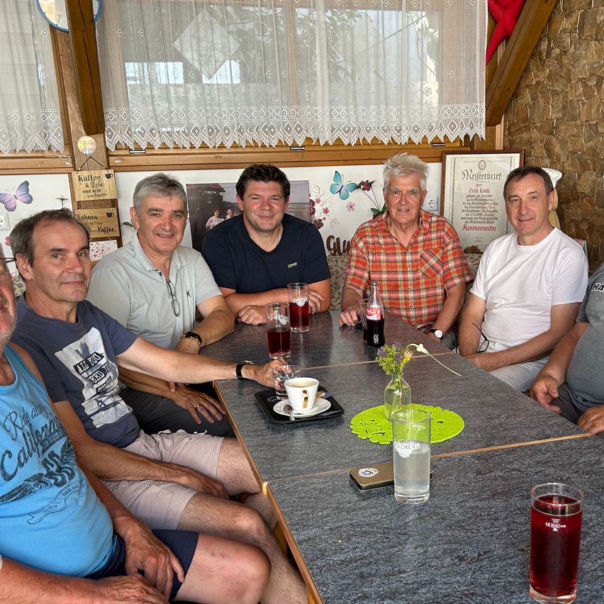 A group of men sits around a table in a restaurant, smiling and posing for a photo. They have drinks and food on the table, and a flower vase is present.